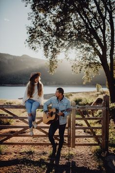 a man holding a guitar next to a woman sitting on top of a wooden fence