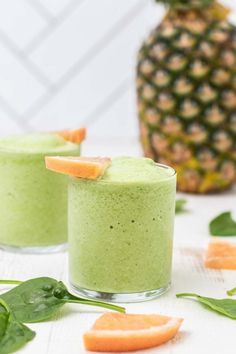 two glasses filled with green smoothie on top of a white table next to sliced oranges