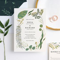 the wedding stationery is surrounded by greenery and gold rings, along with other items