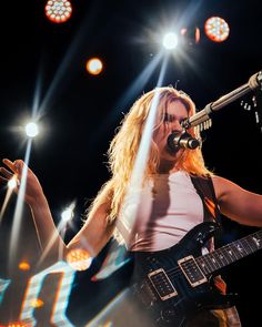 a woman holding a guitar while standing in front of a microphone on stage with lights behind her