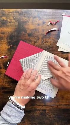 two hands are holding an open book on top of a table with paper and scissors