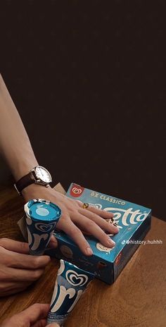 two hands reaching for an unopened cup on top of a wooden table next to a cardboard box