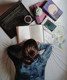 a woman laying on top of a bed reading a book next to a laptop computer