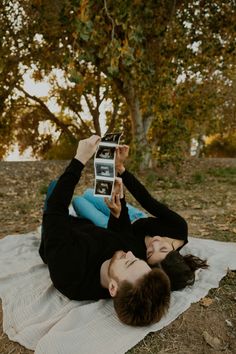 a man laying on top of a blanket next to a tree holding up two photos