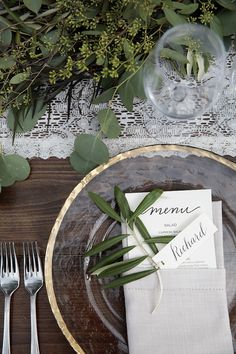 a place setting with silverware, napkins and greenery on a wooden table