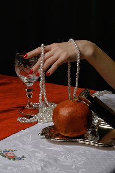 a woman's hand reaching for an apple on a table with pearls and wine glasses