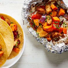 two tacos and a plate of food on a white wooden table with silver foil