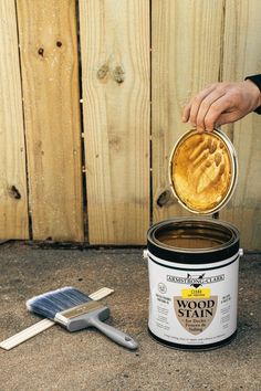 a person holding a paint brush over a can of wood stain next to a wooden fence