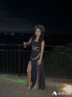 a woman in a black dress standing on a balcony at night wearing a crown and holding a drink