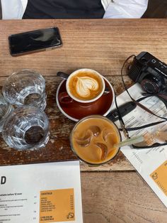 a wooden table topped with two cups of coffee next to glasses and a camera on top of it