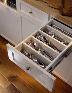 an open drawer with silverware in it on top of a wooden floor next to white cabinets