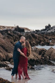 a man and woman standing in the water near some rocks with their arms around each other