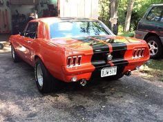 an orange mustang with black stripes parked in front of a garage next to another car