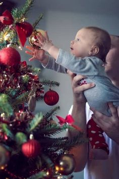 a woman holding a baby next to a christmas tree with ornaments on it and a red ornament hanging from the top