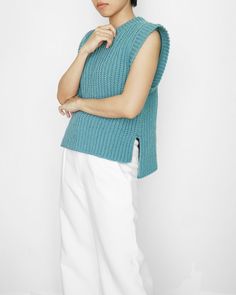 a woman standing in front of a white wall wearing a blue sweater and white pants