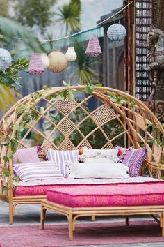 a wicker day bed with pink and white pillows in front of some hanging lanterns