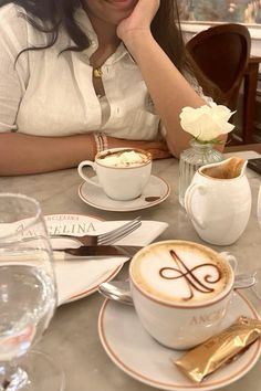 a woman sitting at a table with two cups of cappuccino in front of her