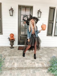 a woman dressed as a witch holding two pumpkins in front of a white house