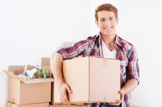 a man holding a cardboard box in front of boxes on top of the floor and smiling