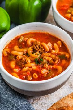 two white bowls filled with pasta soup next to green peppers and pita bread on a table