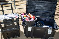 two large black boxes filled with assorted items on top of a dirt ground next to a fence