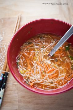 a red bowl filled with noodles and carrots next to chopsticks on a wooden table