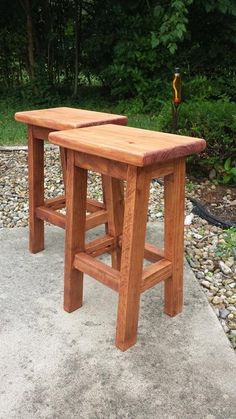 two wooden stools sitting on top of a cement ground next to some bushes and trees