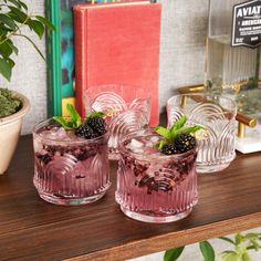 three glasses filled with ice and blackberries on top of a wooden table next to a potted plant