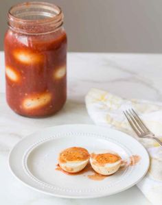 two eggs are sitting on a plate next to a jar of pickled tomatoes and a fork