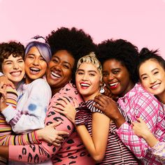 a group of women hugging each other in front of a pink background with one woman smiling