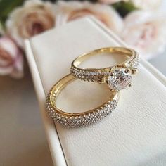 two gold wedding rings sitting on top of a white box with flowers in the background