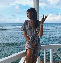 a woman standing on a porch next to the ocean waving at someone in front of her