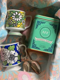 three teacups and two spoons sitting on a floral print table cloth next to a tin of peppermint library