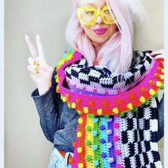 a woman with pink hair wearing glasses and a multicolored scarf holding up her peace sign