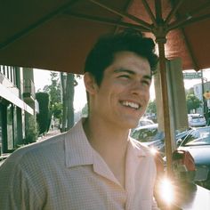 a man smiles under an umbrella on the sidewalk in front of parked cars and buildings