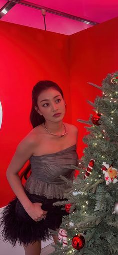 a woman standing next to a christmas tree in front of a red wall with ornaments on it