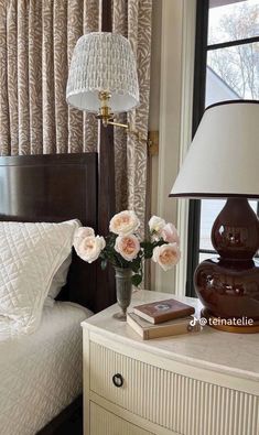 a bedroom with a bed, lamp and flowers on the nightstand next to the window