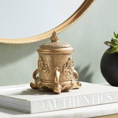 an ornate brass container sits on top of a book next to a mirror and potted plant