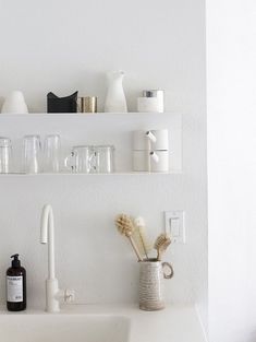 two white shelves above a sink filled with bottles and glasses