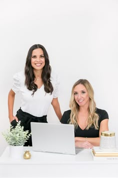 two women sitting at a white desk with a laptop in front of them, both smiling