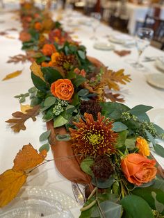 an arrangement of flowers and leaves on a long table with place settings for guests to sit at