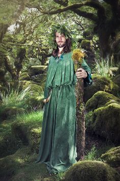 a man dressed in green standing next to a tree with moss growing on the ground