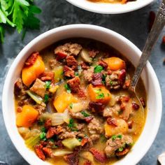 two bowls filled with stew and vegetables on top of a blue tablecloth next to silver spoons