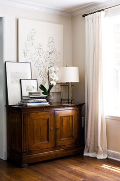 a living room with a wooden cabinet and white curtains on the window sill next to a lamp