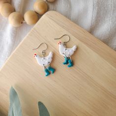 a pair of white and blue rooster earrings sitting on top of a wooden table next to eggs