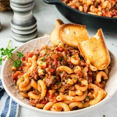 a white bowl filled with pasta and meat sauce next to a slice of bread on the side