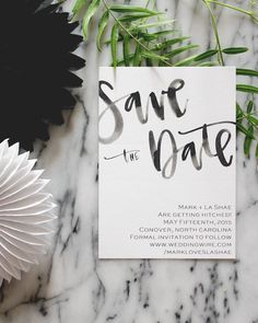 save the date card on top of a marble table with greenery and paper fans