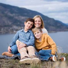 a woman and two boys are sitting on the grass near water with mountains in the background