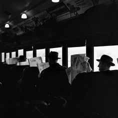 black and white photograph of people reading newspapers on a bus in the dark with lights above them