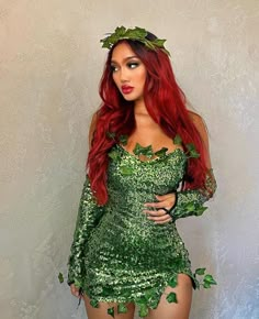 a woman with long red hair wearing a green dress and leaves on her head, standing in front of a wall